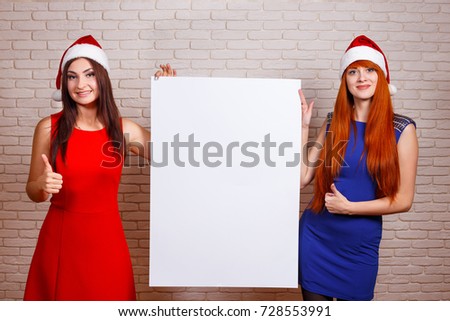 Two beautiful smiling women in Santa caps with thumb up gesture and big empty board in hands. Free space for advertisement or text, mockup for design.New Year, Christmas time sales concept