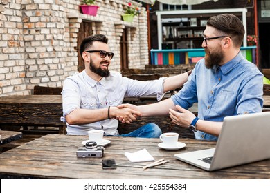 Two beautiful stylish bearded hipster businessman with glasses in a vintage cafe make a deal, shake hands, working on a laptop, tablet, papers, notebook, pens, pencils, drink coffee, mobile phone