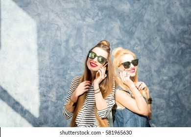 Two beautiful stylish friends using phone on the blue wall background