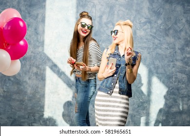 Two beautiful stylish friends using phone on the blue wall background