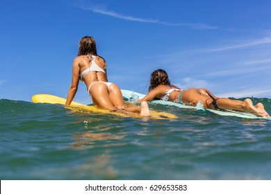 Two beautiful surfing girl with sexy fit butt on surf longboard surfboard board on sunrise or sunset in the ocean. Friends swim to lineup thru waves. Modern active sport lifestyle and summer vacation.
