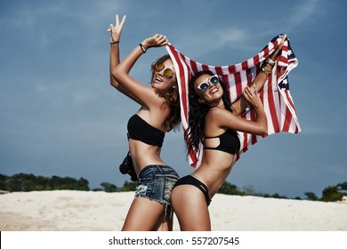 Two beautiful tanned fun hipster girls on the beach, blonde and brunette holding an American flag, old style, laugh, relax on a tropical island, sexy bikini, denim shorts, fashion sunglasses, square