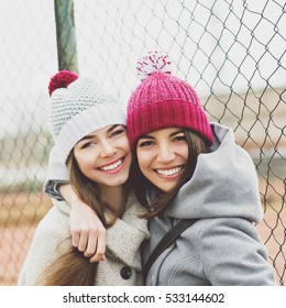 Two Beautiful Teenage Girlfriends Hugging And Smiling Outdoors In Winter, Wearing Coats And Knit Beanie Hats. Two Young Female Friends Portrait Closeup. Retouched, Natural Light.