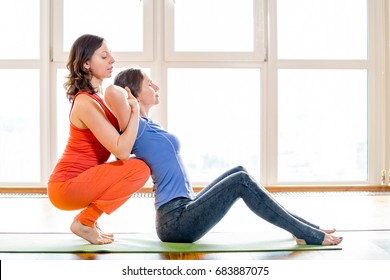 Two beautiful woman in bright colorful sportswear making yoga exercises sitting on mat at yoga studio. Indoor shot. Thai massage element. Treatment, rest, relaxation. Big windows in the background