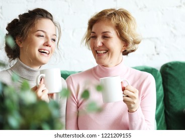 Two Beautiful Women- Mother And Daughter Sitting On The Sofa At Home. They Drink Coffee, Tea.