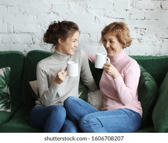 Two Beautiful Women- Mother And Daughter Sitting On The Sofa At Home And Drinking Coffee Or Tea.