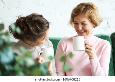 Two Beautiful Women- Mother And Daughter Sitting On The Sofa At Home And Drinking Coffee Or Tea.