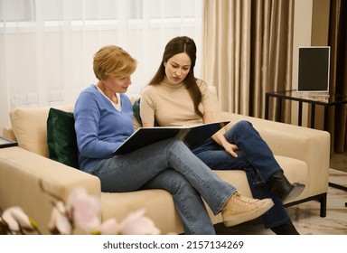 Two Beautiful Women, Mother And Daughter In Travel Agency, Leafing Through A Catalog In A Waiting Room Of A Modern Light Interior Of A Cozy Office Building