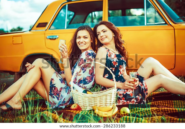 Two beautiful women on the picnic are drinking wine\
near a car.