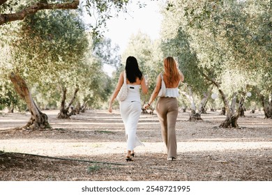 Two beautiful women stroll through a lush olive grove, surrounded by vibrant green trees and sunlight - Powered by Shutterstock