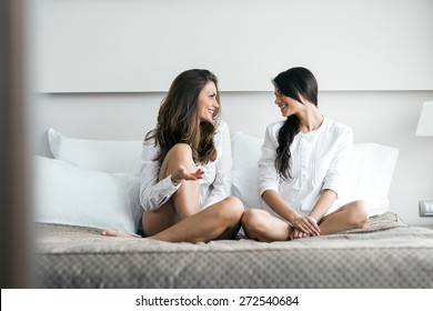 Two beautiful women talking chatting while sitting on a bed of a cozy bedroom
