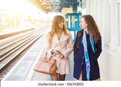 Two beautiful women walking along platform at train station. They are happy, smiling and looking each other. Autumn or winter setting, they are both wearing a coat. Travel and lifestyle concepts.
