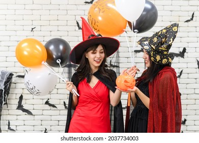 Two beautiful young asian woman in witch halloween costume holding pumpkin Jack O Lantern and balloon on white brick background decorated with black paper bat shape for halloween party - Powered by Shutterstock