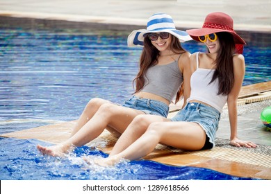 Two Beautiful Young Asian Women In Big Summer Hat And Sunglasses Sitting On The Edge Of The Swimming Pool With Feet In Water Together . Happy Girl Relaxing Kicking Feet Splashing Water.