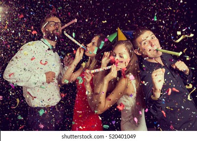 Two beautiful young couples having fun at New Year's party, wearing party hats, dancing and blowing party whistles. Focus on the couple on the right
