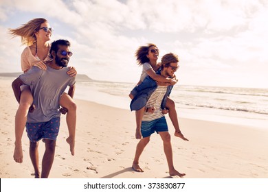 Two beautiful young couples walking by the beach, with men carrying their women on their back. Couples piggybacking on sea shore. Having fun on beach vacation. - Powered by Shutterstock