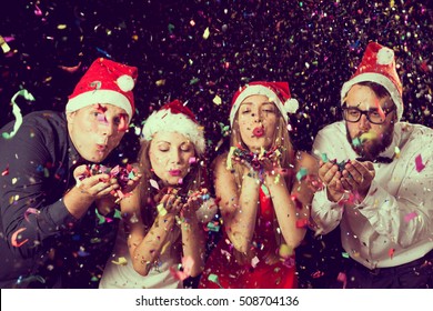 Two beautiful young couples wearing Santa's hats, blowing colorful confetti at midnight at New Year's Eve party - Powered by Shutterstock