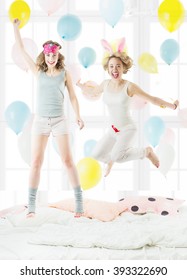 Two beautiful young friends jumping on the bed in sleepwear. pajama party. Cheerful  happy weekend mood. Sunny bedroom with colorful balloons