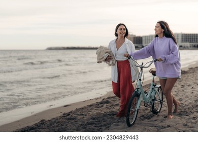 Two beautiful young Latin women in casual clothes with bike walking barefoot on sandy beach. Female tourists on vacation with bicycle and picnic blanket at seashore in the evening. Copy space place. - Powered by Shutterstock