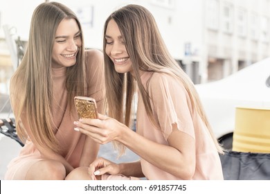 Two beautiful young twins sisters spending time together, looking at mobile phone.