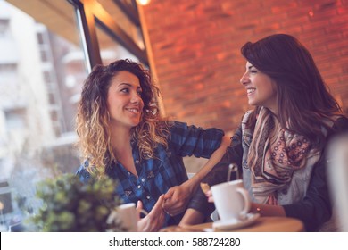 Two Beautiful Young Women Sitting In A Cafe, Drinking Coffee And Having A Pleasant Conversation. Focus On The Girl On The Left