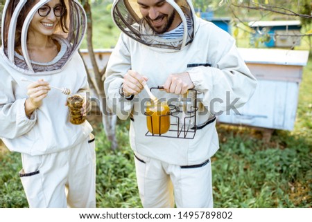 Two beekepers in protective uniform standing together with honey in the jar, tasting fresh product on the apiary outdoors