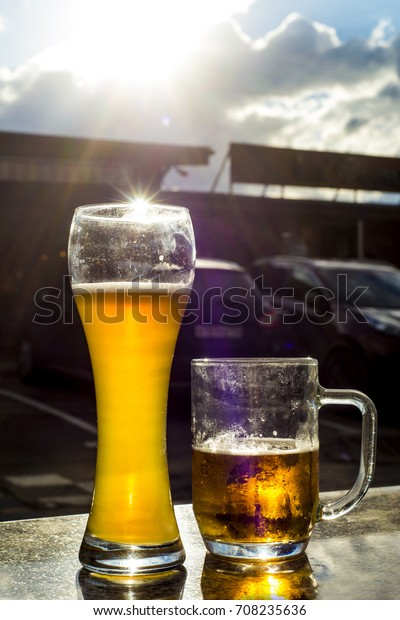Two beer mugs stand on a\
granite table on the background of cars. Drink beer before driving\
a car