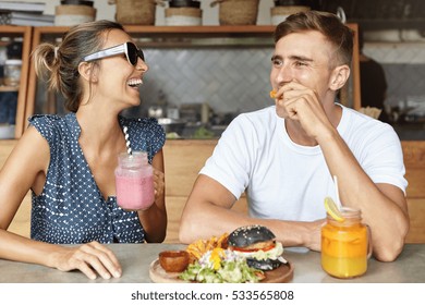 Two best friends having fun together and laughing while eating lunch at coffee shop. Attractive female holding glass of pink smoothie enjoying lively conversation with her handsome boyfriend - Powered by Shutterstock