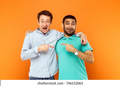 Two best friends hugging, pointing finger each other and looking at camera with shocked face. Studio shot on orange background