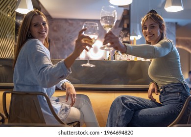 Two Best Friends Toasting At Bar Counter Watching The Camera. Young Women Having Fun Together Raising Wineglasses Sitting At The Restaurant