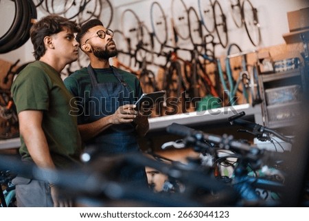 Two bicycle mechanics are using a digital tablet to discuss a repair in a workshop full of bicycles and spare parts