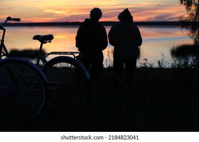 Two Bicycles On The High Shore Of The Lake In Front Of People Looking Ahead At The Sunset At The End Of The Day