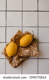 Two Big Lemons With A Green Leaf In A Torn Brown Shopping Craft Paper Bag On A White Tile Kitchen Countertop Background. Minimal Flat Lay With Copy Space. Healthy Immune Support Concept.