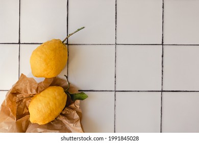 Two Big Lemons With A Green Leaf In A Torn Brown Shopping Craft Paper Bag On A White Tile Kitchen Countertop Background. Minimal Flat Lay With Copy Space. Healthy Immune Support Concept.