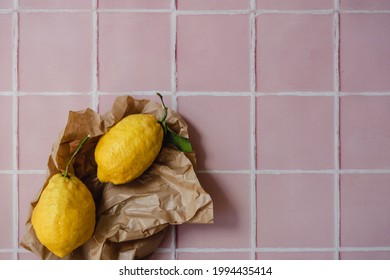 Two Big Lemons With A Green Leaf In A Torn Brown Shopping Craft Paper Bag On A Pink Tile Kitchen Countertop Background. Minimal Flat Lay With Copy Space. Summer Background.