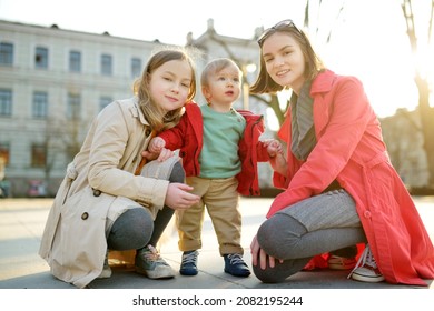 Two Big Sisters And Their Baby Brother Having Fun Outdoors. Two Young Girls Holding Their Baby Boy Sibling On Sunny Spring Day. Kids With Large Age Gap. Kids During City Walk.