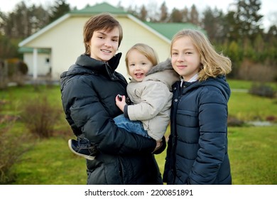 Two Big Sisters And Their Toddler Brother Having Fun Outdoors. Two Young Girls Holding Their Sibling Boy On Winter Day. Kids During Winter Break. Children Exploring Nature Together.