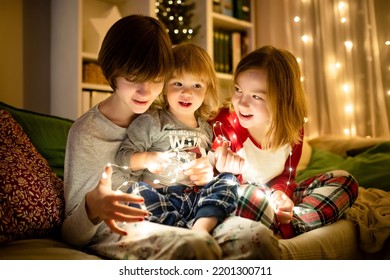 Two big sisters and their toddler brother playing with Chistmas lights in a cozy living room on Christmas eve. Kids spending time at home during winter break. Winter evening with family and kids.