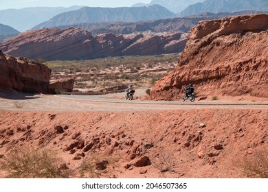 Two Bikers On The Road Through Red Rocky Hills And Mountain