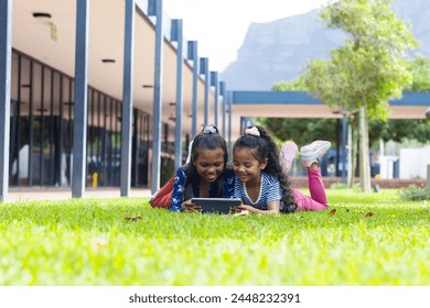 Two biracial girls are engrossed in a tablet outdoors at school, lying on the grass. They enjoy a sunny day at school with a mountain in the background. - Powered by Shutterstock