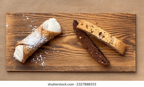 Two biscotti cookies and one cannoli on a wooden plank. Some powdered sugar around the cannoli and some on top. Flat lay photo from above. 