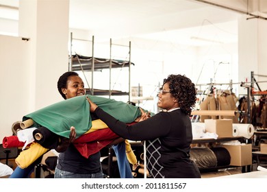 Two black African Zimbabwean woman smiling and laughing working together in a textile factory carrying many different colour rolls of fabric for processing - Powered by Shutterstock