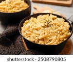 Two black bowls filled with creamy macaroni and cheese, garnished with rosemary, sit on a gray textured surface. Cheese slices on a wooden board are visible in the background a dark cloth.