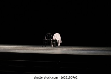 Two Black Chairs Sit On A Black Stage, With Black Background.  One Chair Has A Shawl Draped Over The Back, Top Lit In Theater.