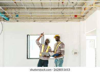 Two Black engineers inspect ceiling infrastructure at a construction site, one pointing upwards while holding a laptop. Image captures teamwork, supervision, and modern building assessment.