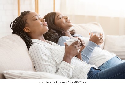 Two black girlfriends resting on sofa with coffee, lying with closed eyes and unwinding after hard day, side view