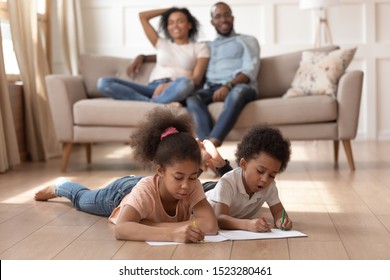 Two black happy kids lying warm heated wooden floor  drawing and markers  smiling blurred parents sitting couch background  Cute children creative art  leisure weekend activities concept 