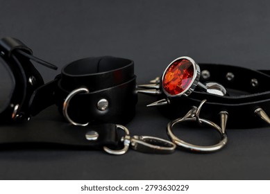 Two black leather collars are placed on a dark surface. One collar has spikes and a large red gem. These collars are meant for pets or as fashion items. They have metal accents and clasps.