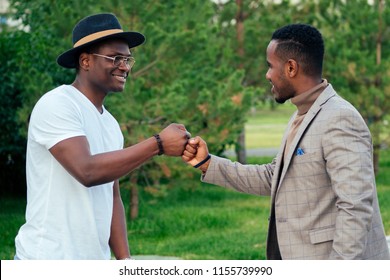 Two Black Men In Stylish Suits Meeting In A Summer Park. African-Americans Friends Hispanic Businessman Embrace Hug Greeting Each Other Teamwork Outdoors. Successful Deal Concept