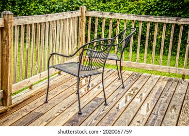 Two Black Metal Lawn Chairs Sitting By Rail On Wooden Deck After The Rain With Lush Greenery In Background